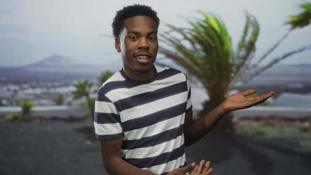 Man in striped shirt shows open palms beside palm tree in front of building under sunlight; optimism.