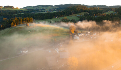Low aerial view over a fog-filled mountain valley with forest, road and scattered houses in soft sunrise light
