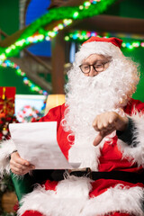 Unhappy actor playing Santa Claus role displeased with letters from kids asking for presents, showing thumbs down. Irked old man disappointed by gift wish lists from children, doing negative gesture