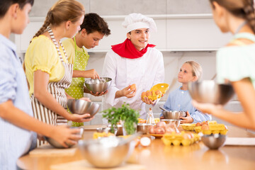 At culinary master class, experienced chef guy in white uniform with egg tray in hands explains subtleties of dessert preparation to children