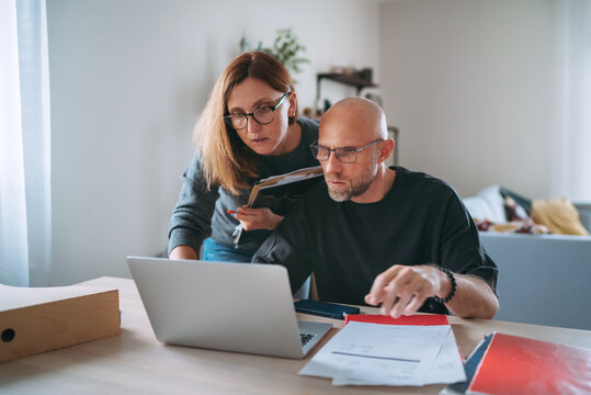 Small business owners focused on financial planning at home office, reviewing paperwork and managing online tasks on laptop, showing teamwork, responsibility and modern entrepreneurial workflow.