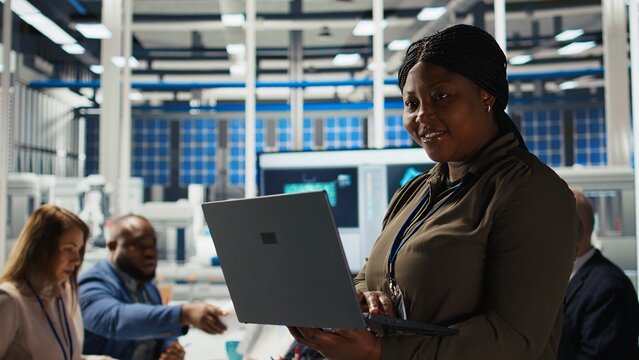 Happy solar panels facility engineer using laptop to improve efficiency. Smiling African american woman working in photovoltaics factory research and development department, using notebook, camera A - Powered by Adobe