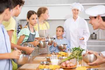 Friendly enthusiastic tween girl learning how to cook and listening to advices of professional chef during group cooking masterclass ..