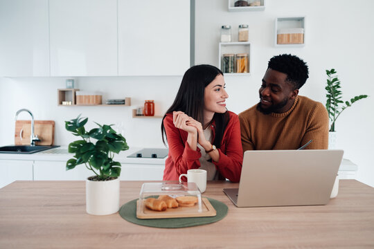 Interracial couple smiling in kitchen using laptop - Powered by Adobe