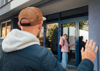 Fototapeta premium Smiling child in warm clothes waves while entering modern school building, creating cheerful morning mood. Dad stands nearby returning gesture highlighting supportive family routine.