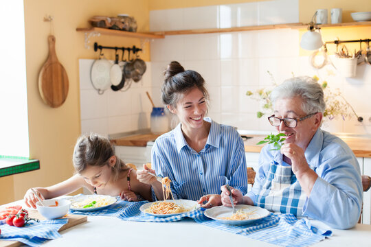 Happy multigenerational family eating pasta together at home kitchen. Three generations enjoying mealtime, healthy food. Woman, child and grandmother have fun. Candid, authentic lifestyle moment