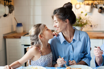 Happy mother and daughter sharing spaghetti and laughing at home kitchen. Family eating pasta, having fun together. Child enjoying homemade dinner, healthy comfort food. Candid, authentic lifestyle