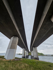 Fototapeta premium A low-angle, centered perspective captures the colossal concrete piers and dual decks of a modern highway bridge spanning a dark, coastal waterway under an overcast sky.