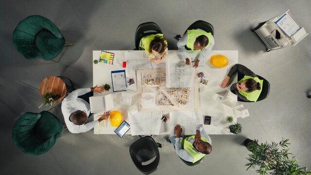 Top down view of property developers and architects in meeting reviewing blueprints and structural designs. Team discusses project approval and city planning for housing growth.