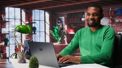 Black remote employee in home office using laptop for web assignments, preparing presentation slides and working on data layouts. Young teleworker finishing company project. Camera B.