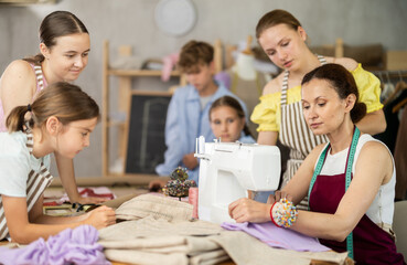 Engaged teenage students, boy and girls, gathering around sewing machine in training workshop while...