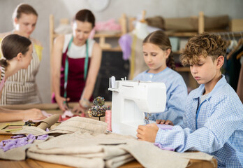 Smiling tween boy focusing on sewing on machine during hands-on lesson while classmates preparing patterns, fabric and fitting garments with female teacher..