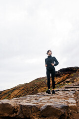 Obraz premium Athletic woman in black activewear stands confidently on rocky terrain, surrounded by natural landscape, embodying strength and determination in outdoor fitness journey