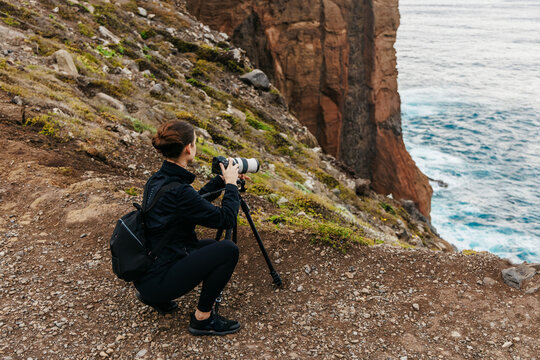 Female photographer capturing stunning coastal landscape with camera on tripod, surrounded by rocky cliffs and ocean waves, showcasing the beauty of nature and adventure