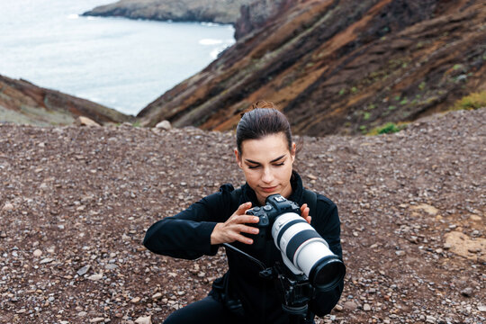 Female photographer adjusting camera settings on a rocky cliff overlooking the ocean, capturing stunning landscapes in a serene travel environment with natural beauty
