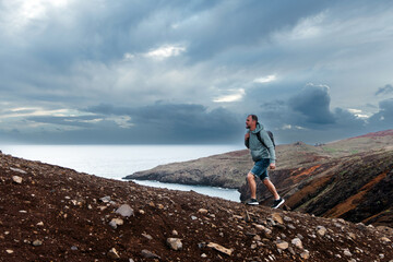 Middle-aged man hiking on rocky terrain near ocean, wearing backpack and enjoying nature, with dramatic clouds and scenic landscape in the background, embodying adventure and exploration