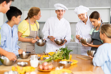 Amiable woman and young man, skilled chefs wearing white cook jackets and toques, running culinary courses for preteen children, sharing secrets of cooking