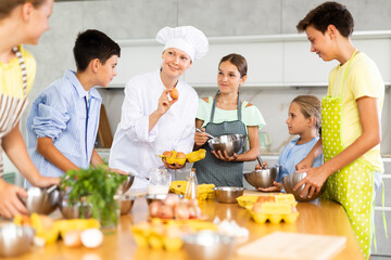 Amiable woman, qualified chef wearing white cook jacket and toque, running culinary courses for preteen children, sharing secrets of cooking ..