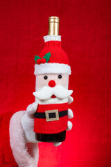 Santa Claus hand holding a wine bottle dressed in a festive Santa Claus-themed knitted cover on red at vertical composition