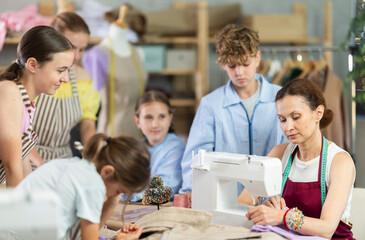 Female teacher demonstrates how to sew on a sewing machine. Children gather around and watch