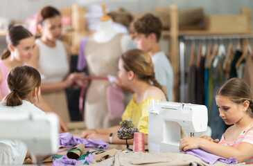 Child works with sewing machine and makes product during lesson at sewing school. Students are actively cooperating with each other in background, discussing completion of assignment.
