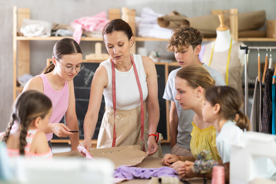 Woman teacher with dress pattern in hands conducts lesson for children at school of cutting and sewing, comments on process of preparing details for creating clothes.