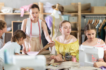 Curious teenage girl attentively watching peer stitching at machine during practical clothing construction course for school students led by experienced sewing instructor in training workshop..