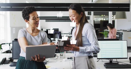 Comparing colleagues in grey tee and blue shirt at open plan office, holding laptop and tablet