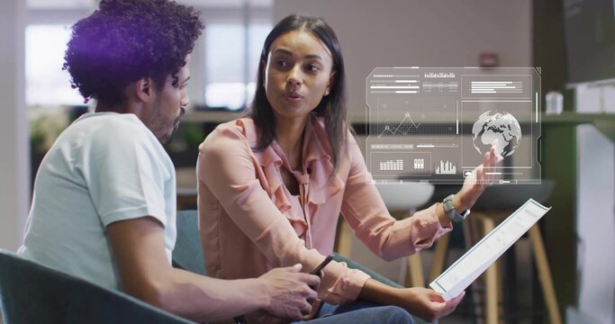 Presenting woman in pink ruffled blouse gesturing at AR data overlay in office, holding tablet