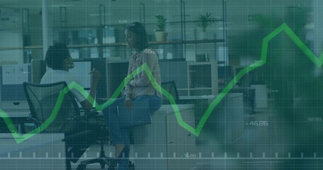 Talking peers in business casual at office with dual screens and laptop showing green graph overlay