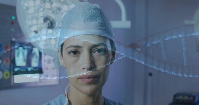 Standing mid-adult female surgeon focusing in OR, wearing cap and scrubs, monitor and DNA overlay