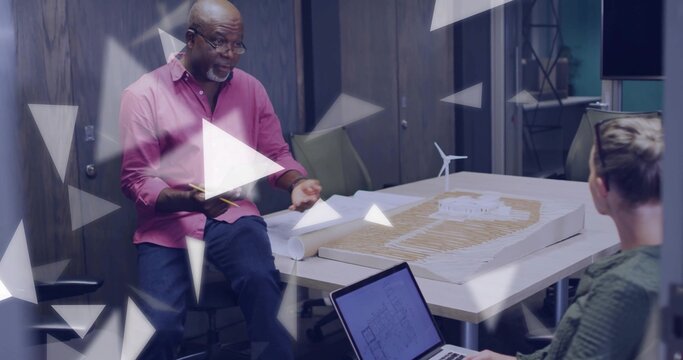 Leading man in pink shirt dark pants discussing scale model in meeting room with laptop, blueprints