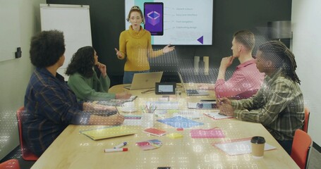 Presenting woman in mustard sweater gesturing at conference table, with laptop and sticky notes