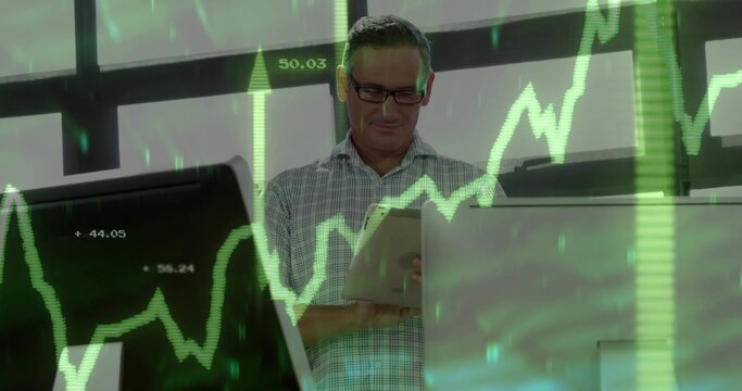 Holding tablet, man in checked shirt scanning green stock charts at desk, with monitors and glasses
