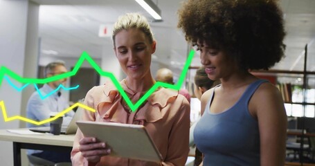 Reviewing tablet, two women wearing peach blouse and blue top in open office with line graphs