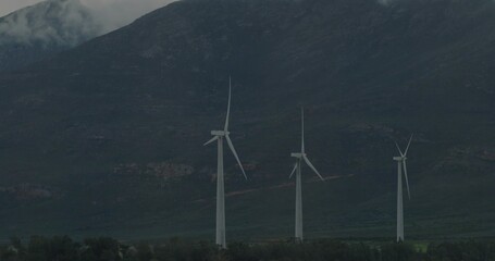 Obraz premium Spinning white wind turbines standing at mountain base, with treeline, rocky slopes, mist