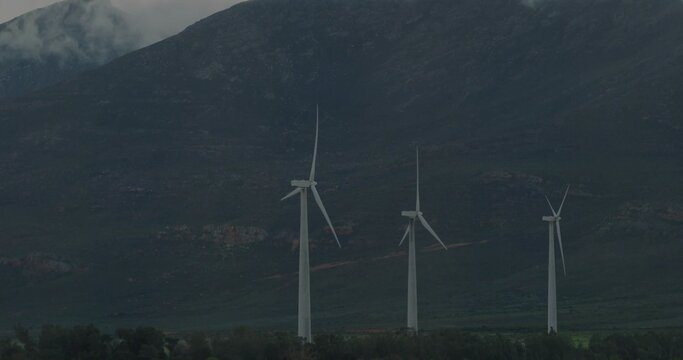 Fototapeta Spinning white wind turbines standing at mountain base, with treeline, rocky slopes, mist