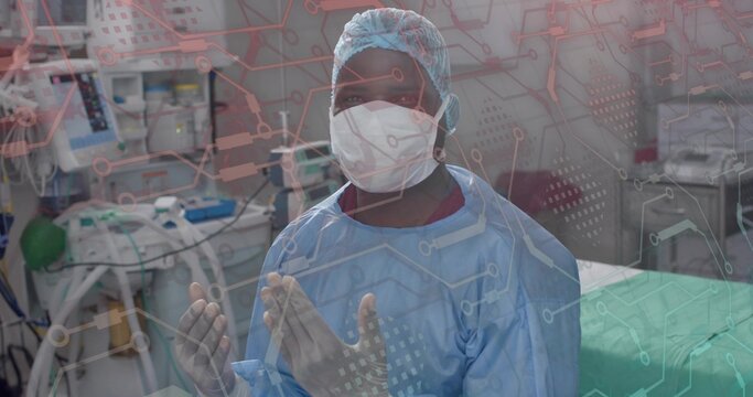 Holding sterile gloves, mid-adult male surgeon in blue gown standing by patient bed with monitors - Powered by Adobe