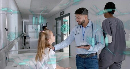 Talking doctor in light-blue shirt kneeling beside child in hospital corridor, holding tablet