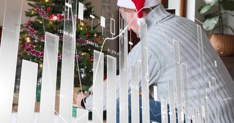 Man in Santa hat kneeling, picking gift and placing it under Xmas tree, finance graphs overlaying - Powered by Adobe