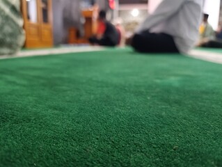 Close up of a vibrant green prayer rug with blurred people praying. Muslim men sitting on a clean green carpet during a worship service. mosque interior during Islamic religious service.