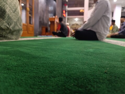 Close up of a vibrant green prayer rug with blurred people praying. Muslim men sitting on a clean green carpet during a worship service. Low angle view of a mosque interior
