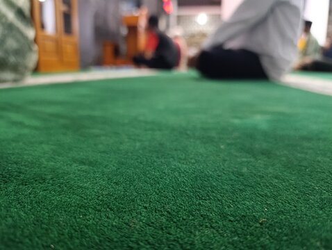 Close up of a vibrant green prayer rug with blurred people praying. Muslim men sitting on a clean green carpet during a worship service. mosque interior during Islamic religious service.