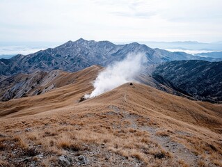 A vast mountain landscape featuring a dry, grassy ridge with a plume of steam rising from a vent. Distant, rugged mountain ranges are visible under a hazy, over