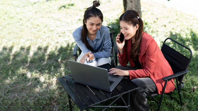 Two Asian women sit outdoors in a sunlit garden; one smiles while on a mobile phone call, looking at a laptop the other woman is pointing at.