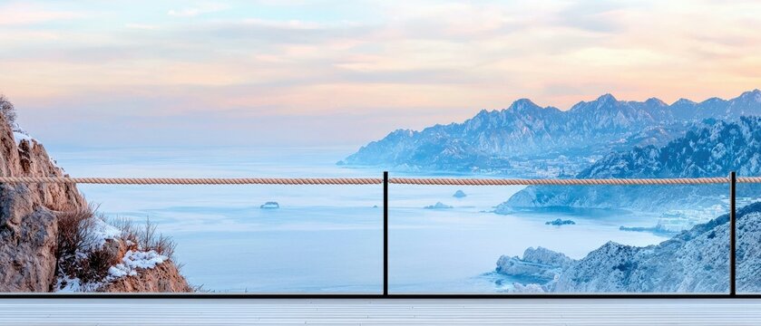 A rope barrier and railing in the foreground, with a vast expanse of calm sea and distant, snow-dusted mountains under a soft, pastel sunrise sky. - Powered by Adobe