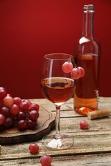 Rose wine in glass, bottle, grapes and corkscrew on wooden table against red background, closeup