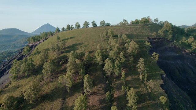 Drone shot showing a sharp boundary between a lush green hill with trees and a steep, dark eroded hillside, with a conical volcano in the distance.

