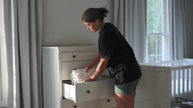 vertical screen white woman stacking nursery linens neatly on dresser top, housekeeper placing folded towels beside crib at bright window, focused service routine with crisp fabrics and soft