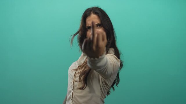 Woman showing middle finger and clenched fist in studio with teal background and intense stare; defiance.